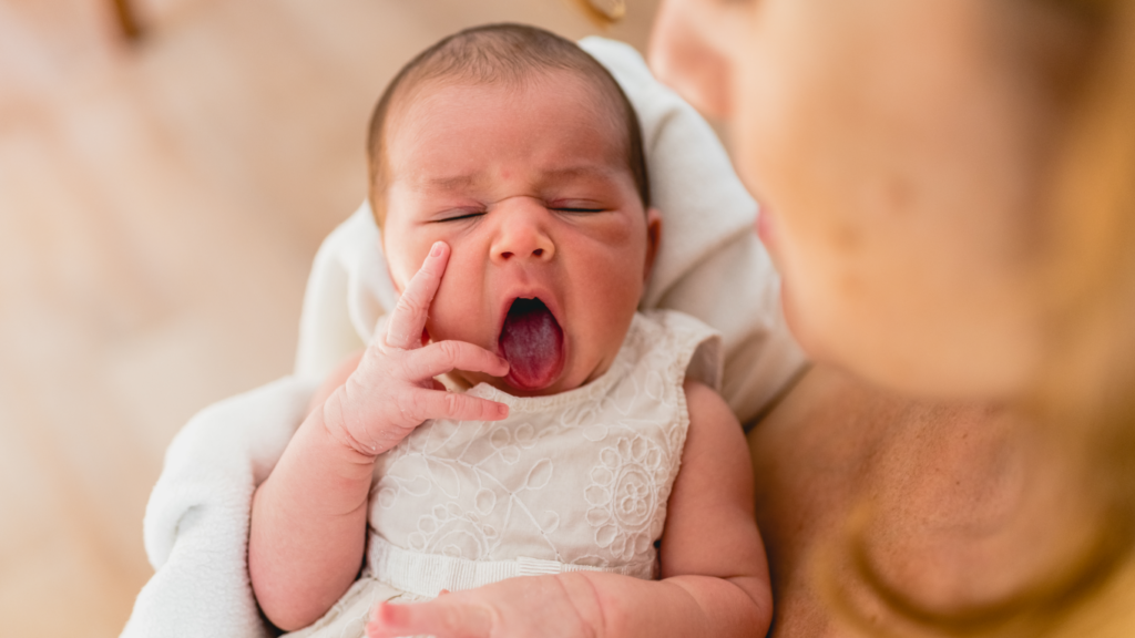 Recém nascida bocejando nos braços da mãe, para falar sobre o teste da linguinha e da orelhinha
